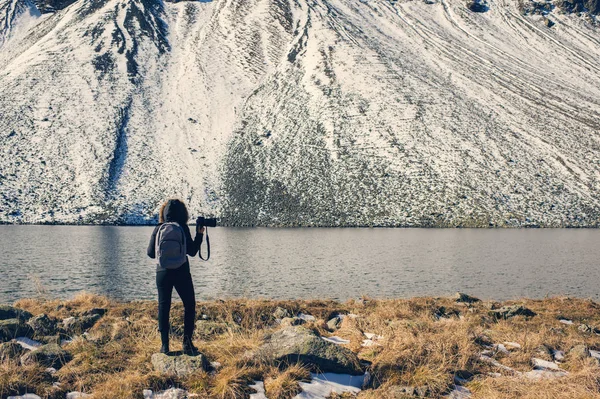 Uma fotógrafa com câmera em uma jaqueta de inverno com pêlo fica em frente às montanhas de neve e um lago na Suíça. Fluela passa na Suíça no inverno . — Fotografia de Stock