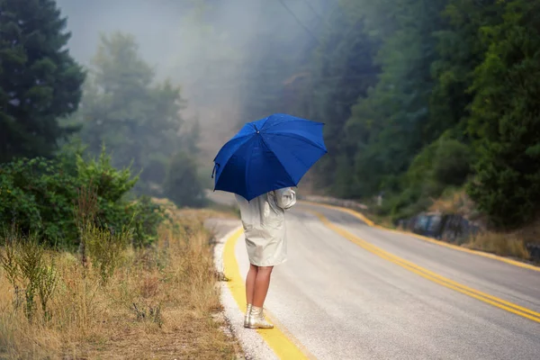 Young female in a raincoat and with umbrella on the road in the fog. Travel of women in the raincoat hitchhiking in the rain