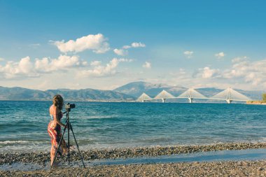 Plajda tripodlu ve kameralı genç bir kadın Rion-Antirion Köprüsü 'nün fotoğrafını çekiyor.