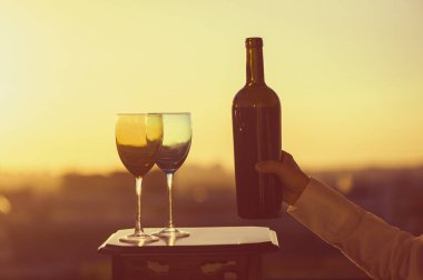 Gün batımında arka planda bir şişe şarap ve iki bardak taşıyan bir kadın eli. Restoranın çatısında servis var.