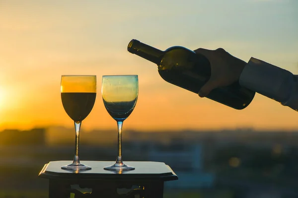Female hand with bottle of wine and two glasses on a sunset background. Service on the roof of the restaurant