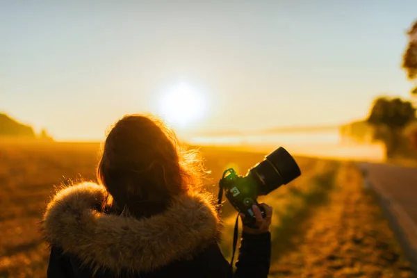 A girl in a jacket with camera on the road against the background of the autumn field and sunset
