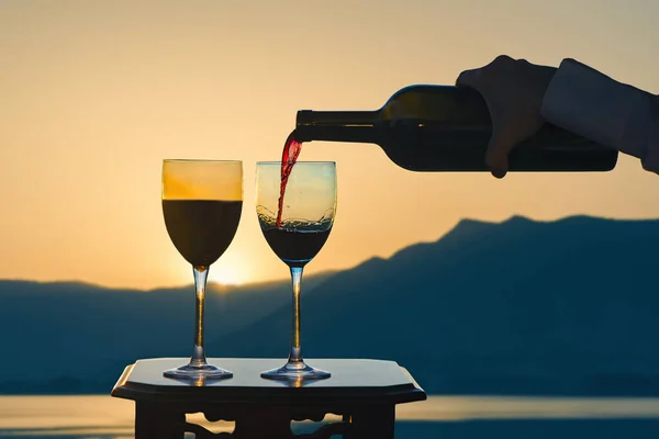 Female hand with bottle pours red wine into glass on lake and mountain background.