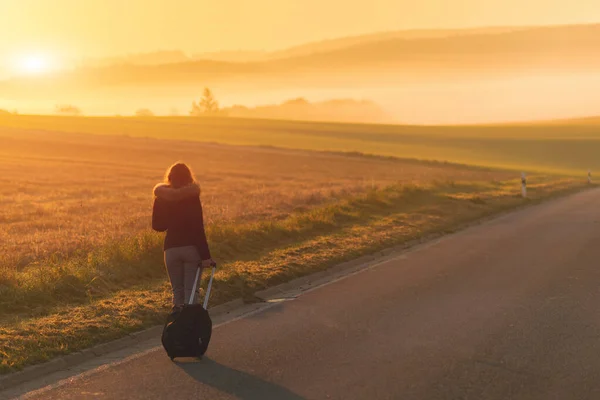 A girl in a jacket with a backpack and suitcase on the road against the background of the autumn field and sunset