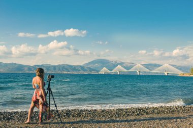 Plajda tripodlu ve kameralı genç bir kadın Rion-Antirion Köprüsü 'nün fotoğrafını çekiyor.