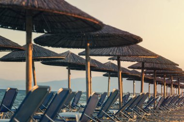 Büyük kamış şemsiyeli tropikal plaj manzarası ve günbatımı deniz arka planında güneşlenme yerleri.