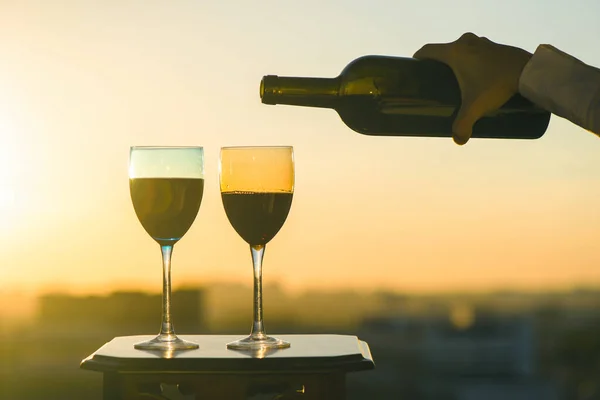 Female hand with bottle of wine and two glasses on a sunset background. Service on the roof of the restaurant