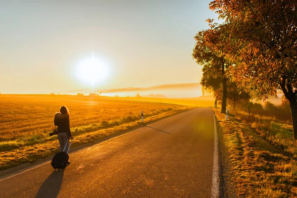 A girl in a jacket with a backpack and suitcase on the road against the background of the autumn field and sunset