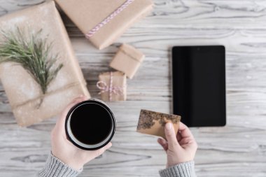 Kahve, kredi kartı ve tableti olan bir kadın. Hediye paketliyorum. Kırsal ahşap arka planda el işi kağıdında karton kutu. Noel ve Yeni Yıl kavramı. 