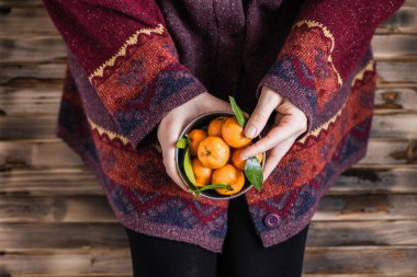 Kocaman bir kazak giymiş bir kadın ahşap kırsal zeminde küçük demir bir kova mandalinayla oturuyor.