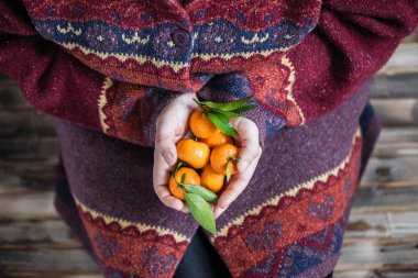 Kocaman bir kazak giymiş bir kadın elinde bir avuç mandalinayla ahşap kırsal zeminde oturuyor.