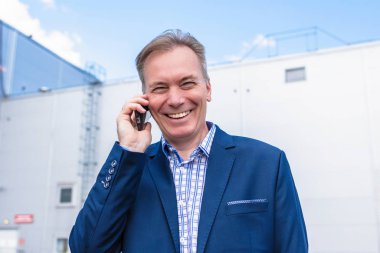 Mavi takım elbiseli orta yaşlı bir işadamı büyük bir fabrikanın özel otoparkında arabasından telefonla ararken gülüyor..