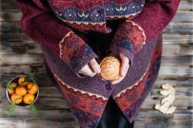 Büyük bir kazak giyen kadın ahşap kırsal zeminde oturur ve mandalinayı temizler.