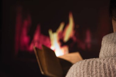 Kışlık kazaklı bir kadın Noel şöminesinin yanında oturmuş bir fincan sıcak içecek ve yün battaniyeyle kitap okuyor. Kış ve Noel tatili kavramı
