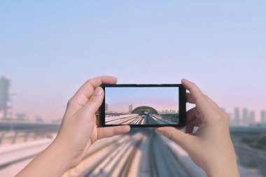 Kadın eli cep telefonuyla Dubai raylarının ve gökdelenlerin fotoğrafını çekiyor. Dubai şehir merkezindeki metro yolunun resmi akıllı telefondan. Birleşik Arap Emirlikleri