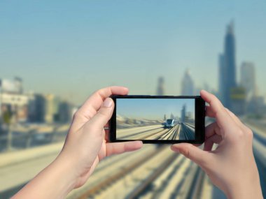 Kadın eli cep telefonuyla Dubai raylarının ve gökdelenlerin fotoğrafını çekiyor. Şehir merkezindeki Dubai metro ve metro treninin resmi. Birleşik Arap Emirlikleri