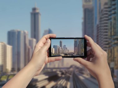 Kadın eli cep telefonuyla Dubai raylarının ve gökdelenlerin fotoğrafını çekiyor. Dubai şehir merkezindeki metro yolunun resmi akıllı telefondan. Birleşik Arap Emirlikleri