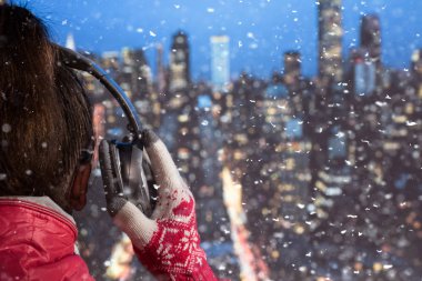 Kışlık ceketli ve örgü eldivenli genç bir kadın kar yağarken New York binasının çatısında kulaklıkla müzik dinliyor. New York şehrinin gökdelenlerinin hava ve manzara manzarası. Noel zamanı Manhattan 'ın göbeğinde