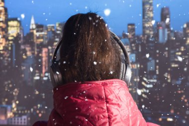 Kışlık ceketli ve örgü eldivenli genç bir kadın kar yağarken New York binasının çatısında kulaklıkla müzik dinliyor. New York şehrinin gökdelenlerinin hava ve manzara manzarası. Noel zamanı Manhattan 'ın göbeğinde