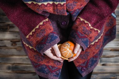 Büyük bir kazak giyen kadın ahşap kırsal zeminde oturur ve mandalinayı temizler.