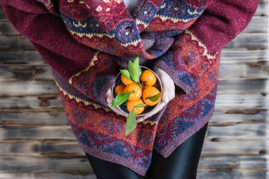 Kocaman bir kazak giymiş bir kadın ahşap kırsal zeminde küçük demir bir kova mandalinayla oturuyor.