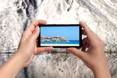 Karlı dağların arka planında, yaz plajının cep telefonundan fotoğraf çeken bir kadının elleri. Mevsimleri değiştirme kavramı