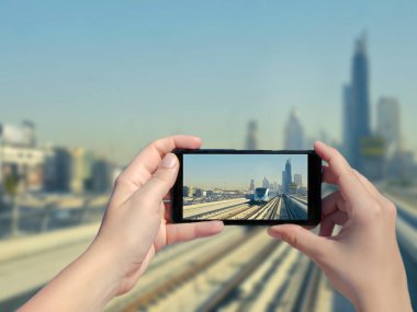 Kadın eli cep telefonuyla Dubai raylarının ve gökdelenlerin fotoğrafını çekiyor. Şehir merkezindeki Dubai metro ve metro treninin resmi. Birleşik Arap Emirlikleri