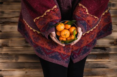 Kocaman bir kazak giymiş bir kadın ahşap kırsal zeminde küçük demir bir kova mandalinayla oturuyor.