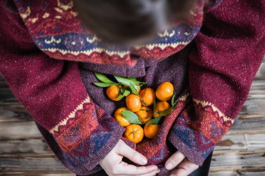 Kocaman bir kazak giymiş bir kadın elinde bir avuç mandalinayla ahşap kırsal zeminde oturuyor.