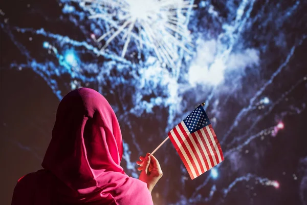 Muslim woman in a scarf holding American flag  during fireworks at night.