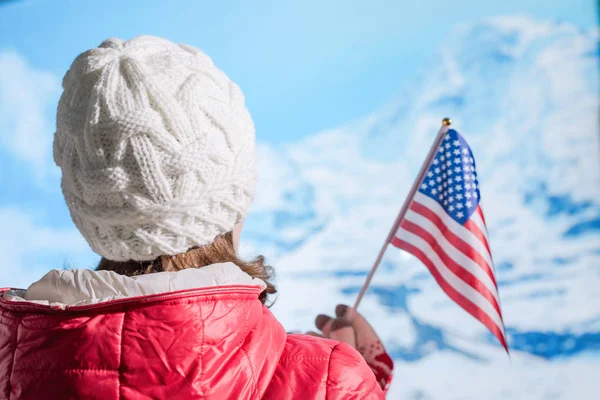 Back view of young woman in a winter knitted white cap, red jacket against the backdrop of the snowy mountains. Girl with american flag on the top of mount