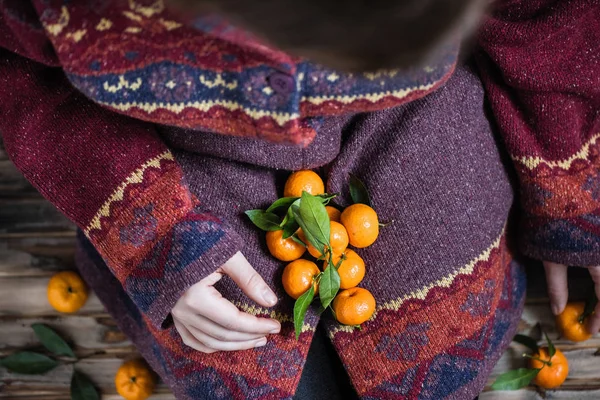 Woman in a huge winter sweater sits on the wooden rustic floor with handful of mandarins in her hands