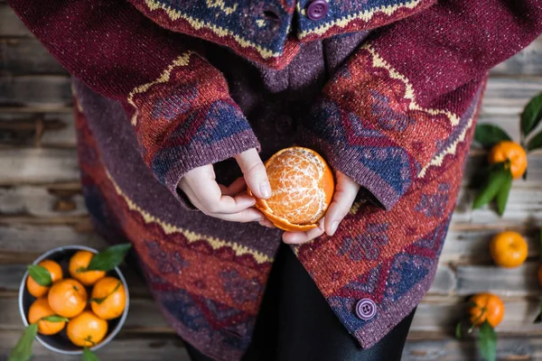 Woman in a huge winter sweater sits on the wooden rustic floor and cleaning the mandarin