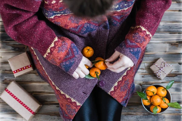 Woman in a huge winter sweater sits on the wooden rustic floor with christmas gifts and mandarins