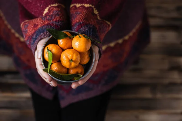 Woman in a huge winter sweater sits on the wooden rustic floor with small iron bucket of tangerines