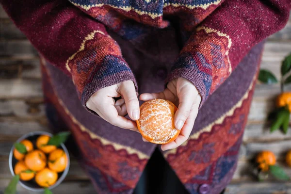 Woman in a huge winter sweater sits on the wooden rustic floor and cleaning the mandarin