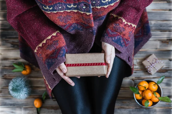 Woman in a huge winter sweater sits on the wooden rustic floor with christmas gifts and mandarins