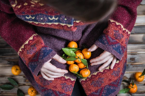 Woman in a huge winter sweater sits on the wooden rustic floor with handful of mandarins in her hands