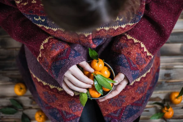 Woman in a huge winter sweater sits on the wooden rustic floor with handful of mandarins in her hands