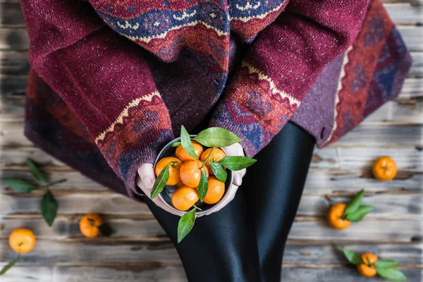 Woman in a huge winter sweater sits on the wooden rustic floor with small iron bucket of tangerines