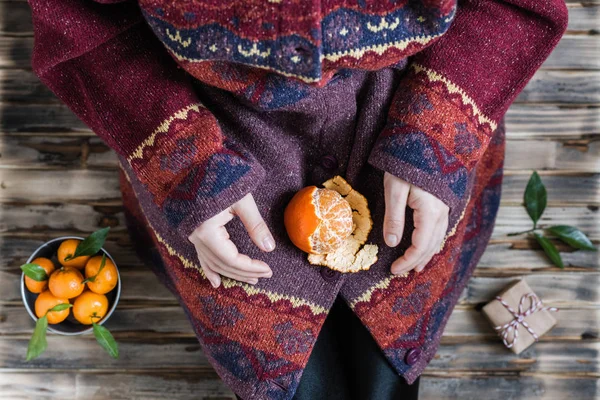 Woman in a huge winter sweater sits on the wooden rustic floor with christmas gifts and mandarins