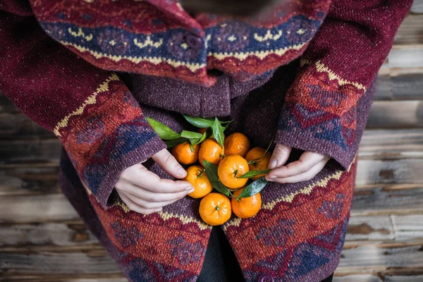 Woman in a huge winter sweater sits on the wooden rustic floor with handful of mandarins in her hands