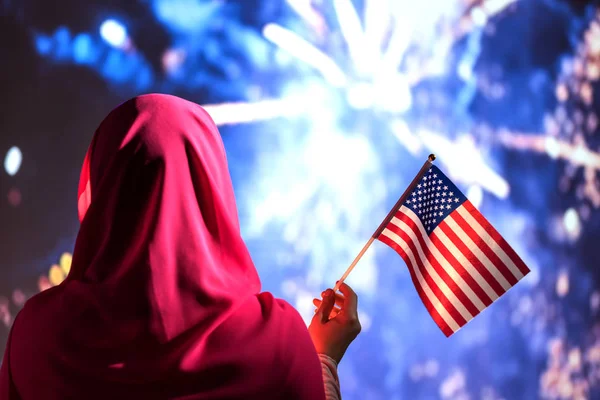 Muslim woman in a scarf holding American flag  during fireworks at night.