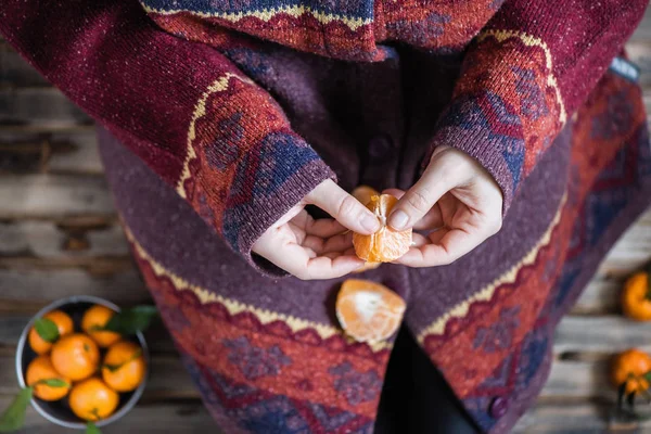 Woman in a huge winter sweater sits on the wooden rustic floor and cleaning the mandarin
