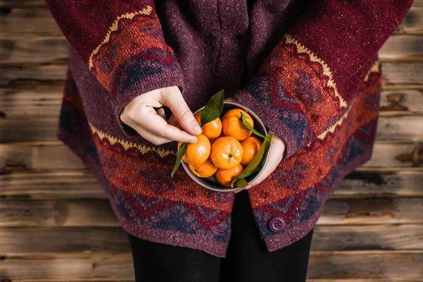 Woman in a huge winter sweater sits on the wooden rustic floor with small iron bucket of tangerines