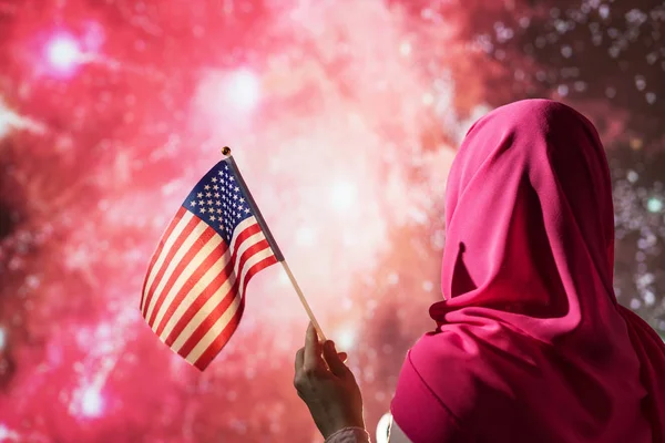 Muslim woman in a scarf holding American flag  during fireworks at night.