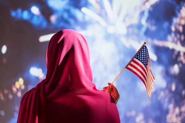 Muslim woman in a scarf holding American flag  during fireworks at night.