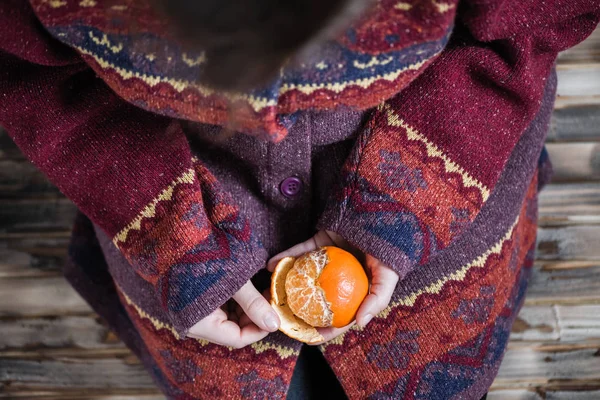 Woman in a huge winter sweater sits on the wooden rustic floor and cleaning the mandarin