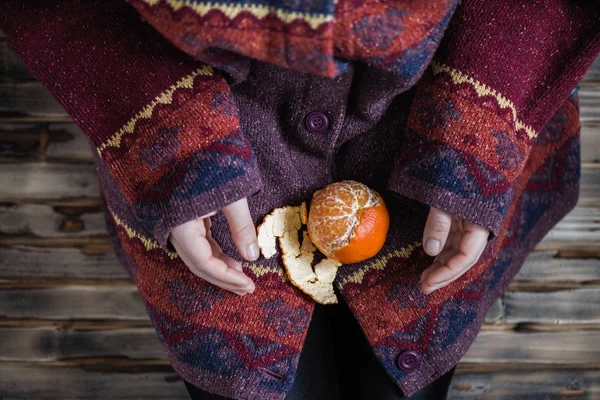 Woman in a huge winter sweater sits on the wooden rustic floor and cleaning the mandarin