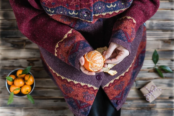 Woman in a huge winter sweater sits on the wooden rustic floor with christmas gifts and mandarins